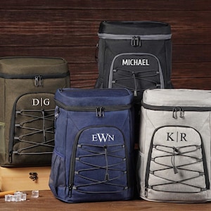 Puede incluir: Cuatro mochilas nevera personalizadas en verde oliva, negro, azul marino y gris. Cada mochila tiene un bolsillo frontal con un diseño de cordón elástico y una cremallera superior. Las mochilas están personalizadas con iniciales o un nombre en blanco.