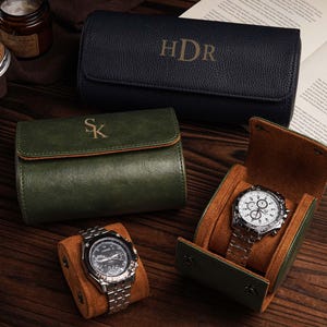 Pu&ograve; includere: Un assortimento di custodie per orologi in pelle verde e blu navy, alcune con iniziali. Sono esposti due orologi con cinturini argentati, uno all'interno di una custodia con interno marrone. Sono visibili anche una candela e un libro.