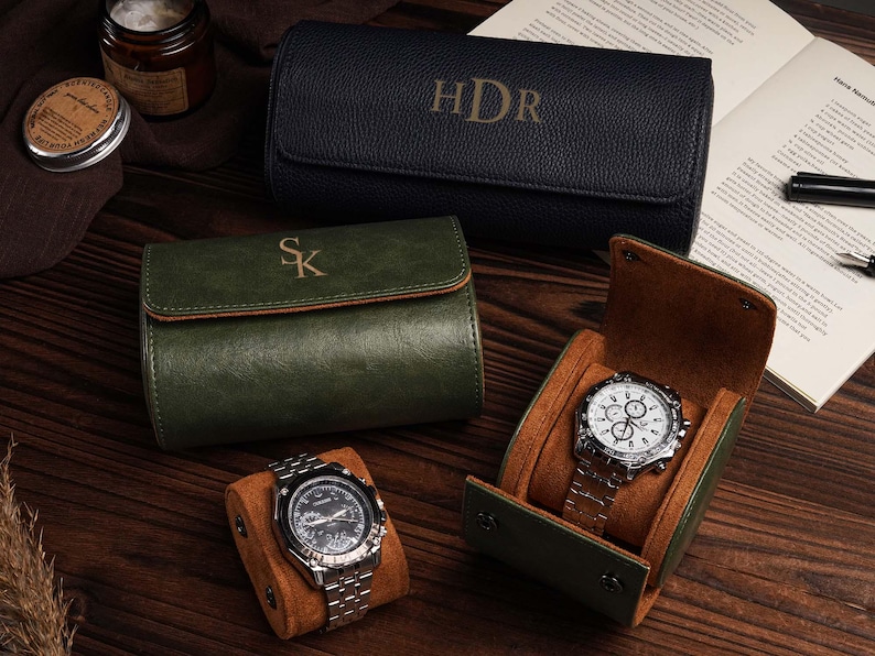 Pu&ograve; includere: Un assortimento di custodie per orologi in pelle verde e blu navy, alcune con iniziali. Sono esposti due orologi con cinturini argentati, uno all'interno di una custodia con interno marrone. Sono visibili anche una candela e un libro.