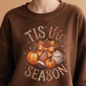 K&ouml;nnte beinhalten: Braunes Sweatshirt mit einem herbstlichen Motiv. Das Design umfasst K&uuml;rbisse, einen Football, eine Tasse Schlagsahne und den Text "Tis the Season". Die Buchstaben haben Karomuster und Leopardenmuster.