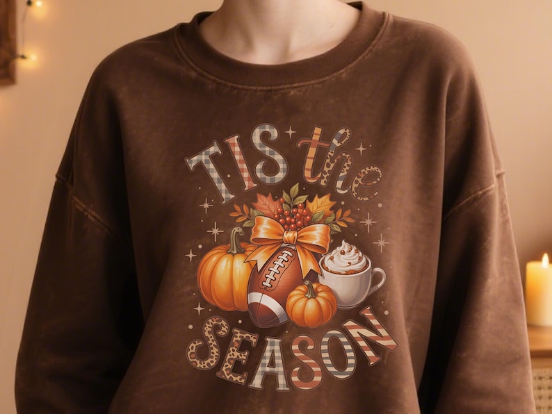 K&ouml;nnte beinhalten: Braunes Sweatshirt mit einem herbstlichen Motiv. Das Design umfasst K&uuml;rbisse, einen Football, eine Tasse Schlagsahne und den Text "Tis the Season". Die Buchstaben haben Karomuster und Leopardenmuster.