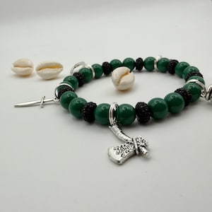 May include: A beaded bracelet featuring dark green and black beads, with silver-toned charms including a small axe and a dagger. The bracelet is accented with small, white seashells. The beads are approximately 0.5 inches in diameter.