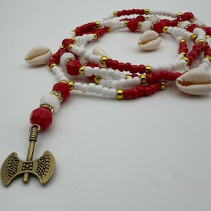 May include: A necklace featuring red and white beads, gold accents, and cowrie shells. A bronze-colored double-bladed axe pendant hangs from the necklace. The beads are strung together in multiple strands.