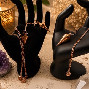 May include: Two copper-toned metal pendulums with pointed tips and matching chains, each draped over a black hand-shaped display stand. A rectangular card is present with the text CASA DE ELEGUA SPIRITUAL GOODS, TAROT & MORE and a sun icon.