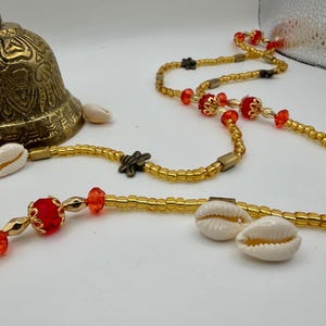 May include: A close-up of a gold-colored bell and beaded necklaces. The necklaces feature gold beads, red accents, and cowrie shells. The bell is ornate with etched designs.