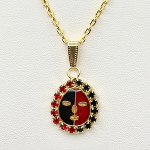 May include: Gold-toned necklace featuring a pendant with a face design. The pendant is divided into black and red sections, framed by red and black gemstones. The chain is a delicate link style.