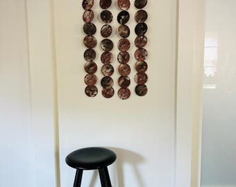 May include: A decorative wall hanging featuring multiple circular elements in shades of brown and copper, suspended from a black bar. A black stool with a round seat and three legs is positioned below the wall art.