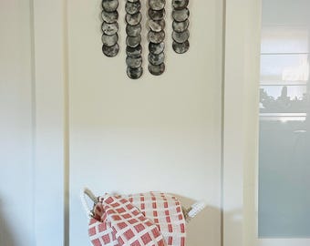 May include: A decorative wall hanging featuring multiple circular, moon-like images suspended from a black bar. Below, a white woven basket holds a red and white checkered throw blanket with fringe.