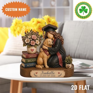 Puede incluir: Figura de graduación que representa a una graduada abrazando a un golden retriever. La figura incluye libros con las palabras "DREAM, BELIEVE, ACHIEVE" y una placa con el nombre "Isabelle CLASS OF 2026". También se incluye un arreglo floral y una pequeña planta.