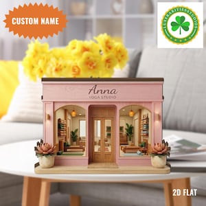 May include: A miniature pink yoga studio model with the name "Anna" above the door. The model has arched windows, a wooden door, and decorative plants. The text "CUSTOM NAME" is in an orange burst shape.