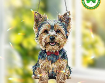May include: A Yorkshire Terrier dog ornament with a red bow tie, suspended by a chain. The dog's fur is a mix of brown, black, and tan. The background is blurred, with green and yellow tones. The ornament is designed for hanging.