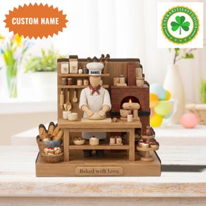 May include: A wooden bakery scene figurine with a chef kneading dough. It features a brick oven, shelves with jars, and a basket of bread. The figurine has the name "Daniel" on the hat and the text "Baked with Love" on the base.
