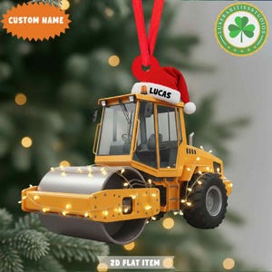 May include: A yellow road roller ornament with a Santa hat that says "LUCAS". The ornament is decorated with string lights and a red ribbon for hanging. The text "CUSTOM NAME" is on a starburst shape.