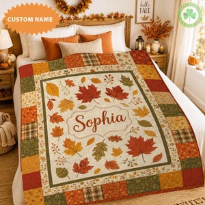 Peut inclure: Une courtepointe personnalisée présentant un panneau central crème avec un motif de feuilles d'automne dans des tons de rouge, orange et vert, avec le nom Sophia imprimé au centre. La bordure se compose d'un patchwork de tissus à carreaux et floraux.