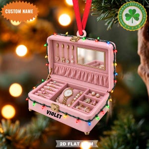 May include: A pink jewelry box ornament featuring a red ribbon hanger and a string of multicolored Christmas lights. The box is open, revealing a mirror, earring hooks, and compartments containing a watch, bracelet, and rings. The name VIOLET is printed on the front.