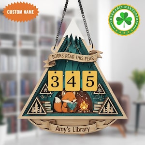 Könnte beinhalten: Holzschild mit Berg- und Waldmotiv, das einen Fuchs zeigt, der ein Buch am Lagerfeuer liest. Das Schild zeigt den Text "BOOKS READ THIS YEAR" und hat Zahlenfelder. Das untere Banner lautet "Amy's Library".