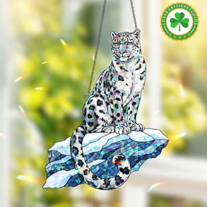 May include: A decorative hanging snow leopard figurine. The figurine is white with black spots and colourful accents, perched on a blue and white geometric ice formation. It hangs from a silver chain. The logo "Luxarities Studios" is in the upper right corner.