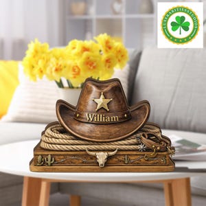 Puede incluir: Un sombrero de vaquero marrón con una estrella dorada y el nombre "William" se encuentra sobre una caja de madera con una cuerda, una calavera de toro y diseños de cactus. El sombrero mide aproximadamente 25 cm de ancho.