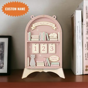 Könnte beinhalten: Ein rosa Bücherregal-Organizer aus Holz mit dem Text „BOOKS READ THIS YEAR“ und „SOPHIE'S LIBRARY“. Es verfügt über Miniaturbücher, eine Katzenfigur und nummerierte Kalenderkacheln. Die Oberseite hat ein individuelles Namensschild.