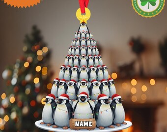 May include: A 2D flat Christmas ornament shaped like a tree, composed of penguins wearing Santa hats. The ornament has a red ribbon for hanging and a yellow star at the top. The text "CUSTOM NAME" is at the top left.