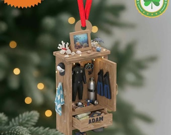 May include: A wooden diving-themed ornament with a wetsuit, fins, and scuba tank inside a storage unit. The drawer is labelled "ADAM". A red ribbon is attached for hanging. The ornament includes a small framed picture and a hat.
