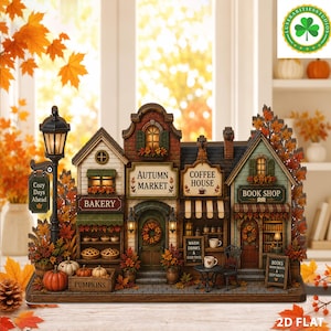Op de afbeelding: Een miniatuur herfstdorp met een bakkerij, herfstmarkt, koffiehuis en boekenwinkel. De structuur bevat een lantaarnpaal met een bordje Cozy Days Ahead, een kist met pompoenen en warme verlichting in alle winkels.