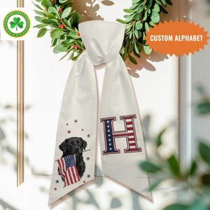 Puede incluir: Bufanda de satén blanco con un Labrador Retriever negro sosteniendo una bandera estadounidense y la letra "H" en rojo y azul. La bufanda tiene un tema patriótico con acentos de estrellas y el texto "CUSTOM ALPHABET".