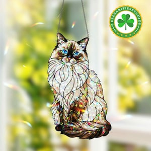May include: A stained glass cat suncatcher with a chain for hanging. The cat has blue eyes, a fluffy white and brown coat, and is sitting upright. The suncatcher has a colorful, iridescent finish. The logo "LUXERARITIESTUDIOS" is in the upper right corner.
