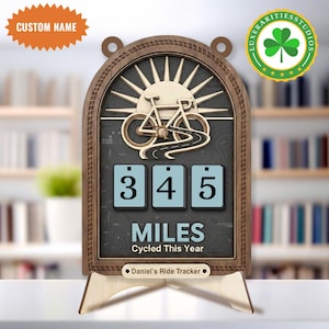 May include: Wooden bicycle ride tracker with a sun and road design. The tracker displays "345 MILES Cycled This Year." Includes a custom name option and a stand. The frame is brown, with a black background and light blue number blocks.