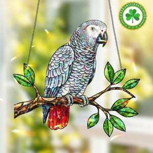 Puede incluir: Un atrapasoles decorativo de loro. El ave está posada en una rama con hojas verdes. El loro tiene plumas grises y blancas, un pico negro y plumas de la cola rojas. El atrapasoles cuelga de una cadena.