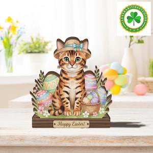 May include: Wooden Easter decoration featuring a tabby cat wearing a straw hat with a blue bow. The cat sits in front of decorated Easter eggs, a basket of eggs, and the words "Happy Easter!" on a wooden base. The cat has green eyes.
