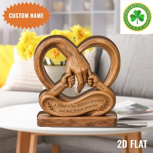 Puede incluir: Una escultura de madera con un diseño en forma de corazón, que representa la mano de un padre sosteniendo dos manos más pequeñas. Los nombres "Sophia" y "Alex" están inscritos. El texto dice: "Un padre es el primer héroe de sus hijos y su protector para siempre."