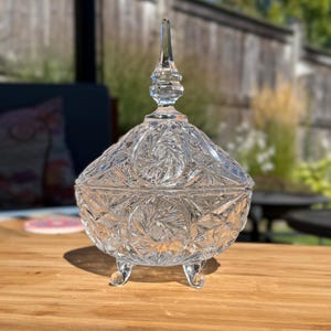 May include: A clear, cut-glass candy dish with a lid, resting on three small feet. The dish features intricate starburst and floral patterns, and a tall, pointed finial on the lid. The dish is displayed on a wooden surface.