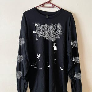 Op de afbeelding: Zwart shirt met lange mouwen en een wit grafisch ontwerp. Het ontwerp bevat een sleutel, geometrische vormen en figuren. De mouwen hebben extra sleutel- en geometrische ontwerpen.