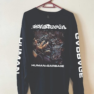May include: A black long-sleeve shirt featuring a graphic print of a hand reaching out from a pile of scrap metal. The text DYSTOPIA appears above the image, and HUMAN=GARBAGE is printed below. The word HUMAN is printed vertically on both sleeves.