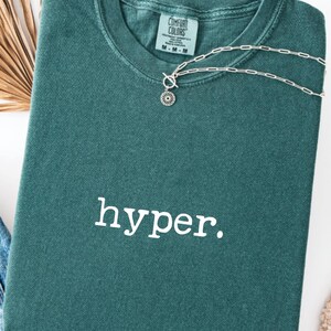 May include: A folded teal Comfort Colors t-shirt with the word "hyper." printed in white. A silver necklace with a floral pendant rests on the shirt. The shirt has a crew neck and a tag that reads "M-M-M".