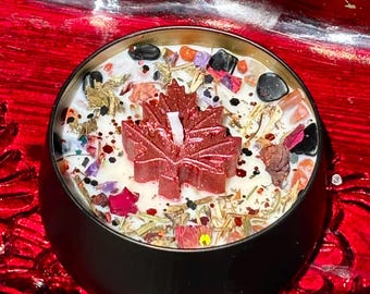 Op de afbeelding: Een zwart metalen kaarsblik gevuld met witte was, bekroond met een donkerrode esdoornblad-vormige wax melt, verspreide zwarte stenen, gedroogde bloemblaadjes, kleine oranje kristallen en fijne glitterdeeltjes op het oppervlak.