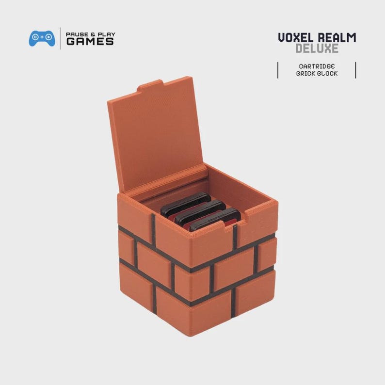 May include: A brick-shaped storage box with an open lid, holding three black game cartridges. The box is made of orange-brown material with black lines to simulate bricks. The text "VOXEL REALM DELUXE" and "CARTRIDGE BRICK BLOCK" are visible.