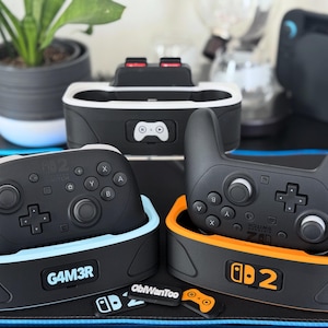 May include: Black and gray gaming controllers and charging docks. The controllers have buttons labeled X, Y, A, and B. The charging docks are black with blue and orange accents. The text "G4M3R" and "ObiWanToo" are visible.
