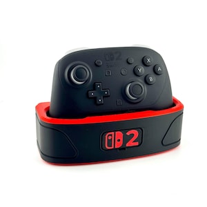 May include: A black Nintendo Switch Pro controller with white accents, resting in a red and black charging dock. The controller has buttons labeled X, Y, A, and B. The dock has the Nintendo Switch logo.