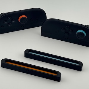 May include: Black game controller accessories with orange and blue accents. The image shows two controllers and two matching rails. The controllers have buttons and joysticks. The rails have colored stripes.