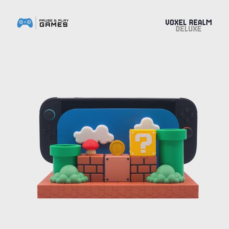 May include: A black handheld gaming console with a blue screen displaying a cartoon scene with white clouds, green pipes, and a yellow question mark block. The text "VOXEL REALM DELUXE" and "PAUSE & PLAY GAMES" are visible.