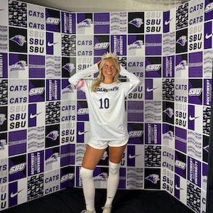 May include: A person wearing a white long-sleeved jersey with the number 10, white shorts, and knee-high socks. The jersey has "Bearcats" printed on it. The background is a wall with a repeating pattern of "SBU" and "Bearcats" logos.