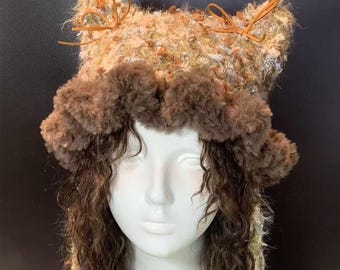 May include: A unique, handmade hat with a textured, multi-colored crown and a fluffy, brown brim. The hat features small, decorative leather bows and is designed to be worn on the head. The hat is displayed on a mannequin head.