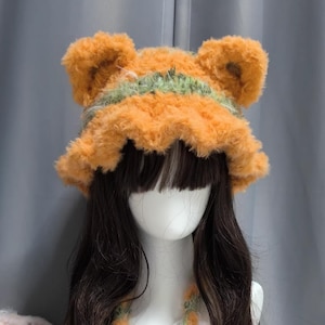 Puede incluir: Un sombrero naranja esponjoso con orejas de oso y una franja verde. El sombrero está hecho de un material suave y texturizado. El sombrero está diseñado para ser usado en la cabeza. Un accesorio divertido y caprichoso.