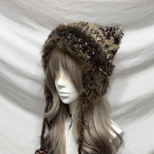 May include: A brown, furry hat with pointed ears and long braided tassels. The hat features a mix of textures and colors, including dark brown, beige, and green. The hat is designed to be worn on the head and has a unique, whimsical style.