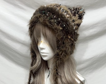 May include: A brown, furry hat with pointed ears and long braided tassels. The hat features a mix of textures and colors, including dark brown, beige, and green. The hat is designed to be worn on the head and has a unique, whimsical style.