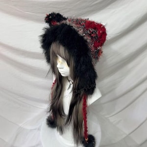 Puede incluir: Una capucha de piel sintética negra con detalles rojos y plateados, con orejas adjuntas y largas hebras trenzadas con pompones. La capucha se muestra en una cabeza de maniquí con cabello largo y oscuro.