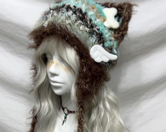 May include: A whimsical, handmade hat with cat ears, featuring a mix of brown, teal, and cream-colored yarn. The hat is adorned with a white bone and a pair of white wings. The hat has long, brown ear flaps.