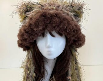 May include: A whimsical, handmade hat with a brown, fluffy brim and matching ear-like accents. The hat is adorned with a mix of green, tan, and black textured materials, creating a unique, textured appearance. Long, flowing strands of similar materials cascade down.